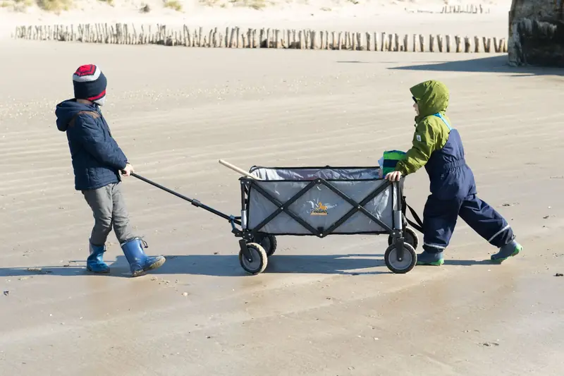 Two children pulling and pushing a grey folding wagon across a sandy beach
