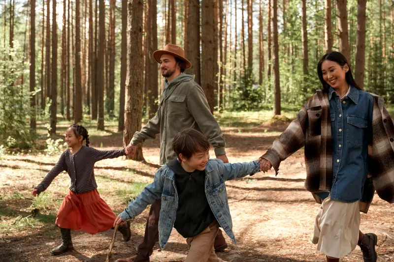 Family of four holding hands and walking together through a sunlit pine forest