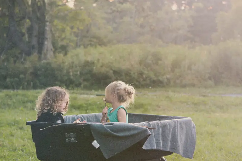 Two young children sitting side by side in a black folding wagon on a sunny lawn